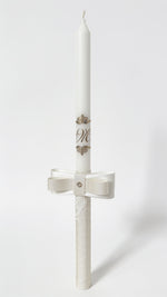 A white christening candle with a matching ecru ribbon bow at the base, decorated with small gold floral motifs and monogram lettering, shown on a light gray background.