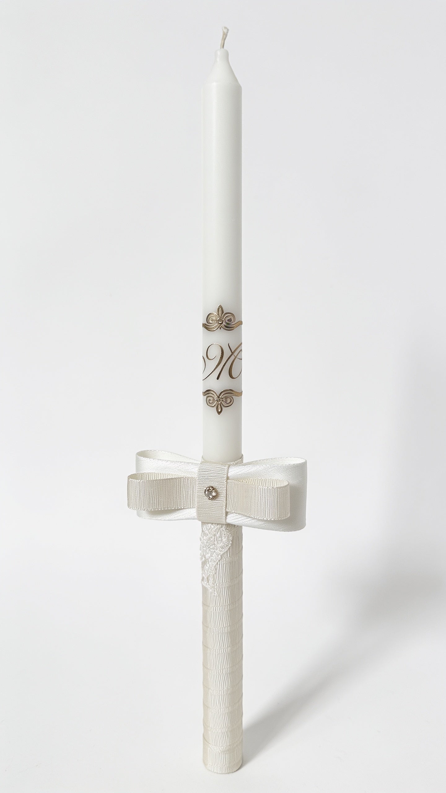 A white christening candle with a matching ecru ribbon bow at the base, decorated with small gold floral motifs and monogram lettering, shown on a light gray background.
